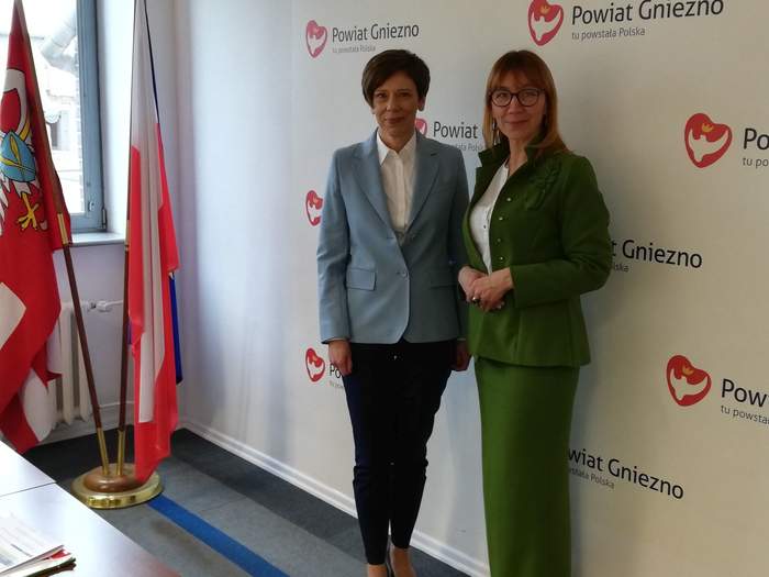 Pokaż zdjęcie: Dwie kobiety pozują do zdjęcia na tle ścianki powiatu gnieźnieńskiego. Przy oknie widoczne są dwie flagi oraz fragment biurka.