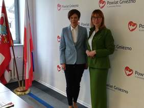 Pokaż zdjęcie: Dwie kobiety pozują do zdjęcia na tle ścianki powiatu gnieźnieńskiego. Przy oknie widoczne są dwie flagi oraz fragment biurka.