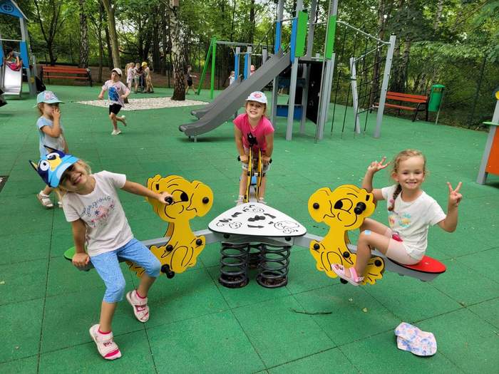 Pokaż zdjęcie: Grupa uśmiechniętych dzieci znajduje się na placu zabaw przy różnego rodzaju urządzeniach. Na placu zabaw widoczne są między innymi: huśtawki, drabinki, zjeżdżalnie, ławka oraz kosz na śmieci.