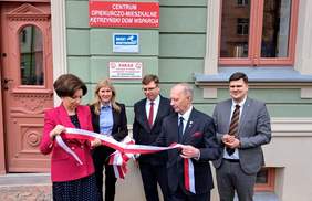 Pokaż zdjęcie: Na pierwszym planie dwie elegancko ubrane osoby trzymają w rękach wstęgę, za nimi trzy osoby na tle budynku