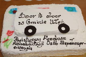 Pokaż zdjęcie: Biały tort w kształcie busa z logami PFRON czyli  pomarańczowy tulipan podparty z zieloną łodygą.Napis na torcie door to door  w kolorze brązowym