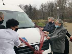 Pokaż zdjęcie: Fragment przodu białego busa z bordową wstążką związaną w kokardę, dwie osoby z jednej strony auta i 3 z drugiej trzymają w rękach nożyczki i przecinają wstęgę.