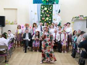 Pokaż zdjęcie: W sali stowarzyszenia trwa występ artystyczny. Na pierwszym planie stoi kobieta, która śpiewa do mikrofonu. Za nią znajduje się chór. Z prawej i lewej strony widać ludzi siedzących za stołami. 