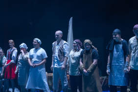 Pokaż zdjęcie: Zdjęcie z bliska, przedstawiające grupę kilkunastu osób w strojach scenicznych, stojących boso na scenie, ułożonych w linii do ukłonu po występie. Przód sceny udekorowany jest niebieskim materiałem imitującym wodę. Po lewej stronie stoją dwie osoby w strojach galowych: kobieta i mężczyzna z mikrofonem.