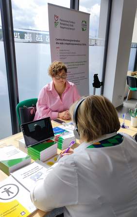 Pokaż zdjęcie: Stoisko informacyjne PFRON. Osoba zainteresowana rozmawia z ekspertem PFRON - rozmowa prowadzona jest online. Na stoliku dostepne są ulotki oraz informatory PFRON.