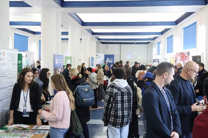 Pokaż zdjęcie: Zdjęcie korytarza, po jego obydwu stronach stoją wystawcy, którzy mają na stolikach dużo ulotek. Pomiędzy stolikami stoją zainteresowane osoby.