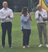 Osoba przemawia do mikrofonu na stadionie podczas turnieju bocci, obok stoją uczestnicy i klaszczą. W tle widoczna dmuchana atrakcja oraz ogrodzenie boiska i drzewa.