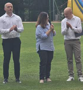 Osoba przemawia do mikrofonu na stadionie podczas turnieju bocci, obok stoją uczestnicy i klaszczą. W tle widoczna dmuchana atrakcja oraz ogrodzenie boiska i drzewa.
