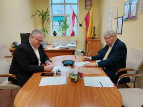 Pokaż zdjęcie: Przy podłużnym stole siedzi naprzeciwko siebie dwóch mężczyzn. Podpisują umowę. Na biurku leżą różne dokumenty, pieczątki, długopisy. 