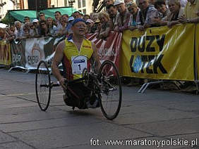 Pokaż zdjęcie: Zawody kolarskie. Na pierwszym planie bohater artykułu klęczy na stopniach roweru ręcznie napędzanego.