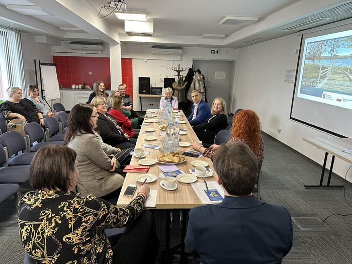 Pokaż zdjęcie: Przy długim drewnianym stole siedzi kilka osób, głównie kobiety. Po prawej wisi ekran z prezentacją. Po lewej stronie krzesła na których siedzą ludzie.