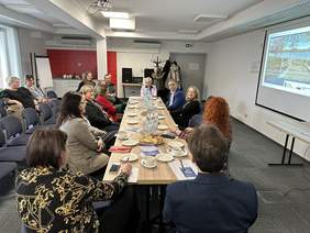 Pokaż zdjęcie: Przy długim drewnianym stole siedzi kilka osób, głównie kobiety. Po prawej wisi ekran z prezentacją. Po lewej stronie krzesła na których siedzą ludzie.