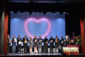 Pokaż zdjęcie: Kilkanaście osób odświętnie ubranych stoi na scenie. Każda z nich trzyma w ręku kwiat i dyplom. W tle na dużym ekranie wyświetlone jest serce.