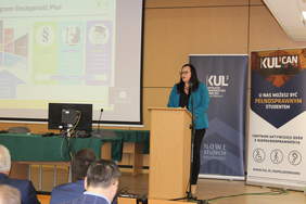 Pokaż zdjęcie: Zdjęcie mównicy robione ze środka auli, przy mównicy stoi kobieta z długimi ciemnymi włosami ubrana w niebieski garnitur