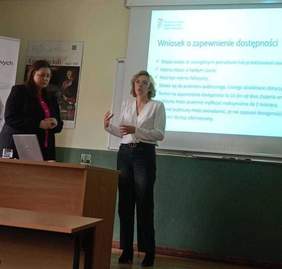 Pokaż zdjęcie: Dwie kobiety stoją w sali konferencyjnej. Jedna z nich przemawia, gestykulując rękoma. Z kolei druga jest zamyślona i ma wzrok skierowany ku podłodze. W tle znajduje się ekran z prezentacją przedstawiającą składanie wniosku o zapewnienie dostępności.