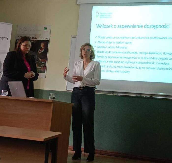 Pokaż zdjęcie: Dwie kobiety stoją w sali konferencyjnej. Jedna z nich przemawia, gestykulując rękoma. Z kolei druga jest zamyślona i ma wzrok skierowany ku podłodze. W tle znajduje się ekran z prezentacją przedstawiającą składanie wniosku o zapewnienie dostępności.