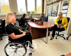 Pokaż zdjęcie: Przy owalnym stole siedzą cztery kobiety. Jedna z pań siedzi na wózku inwalidzkim, a inne na krzesłach. Rozmawiają ze sobą. 