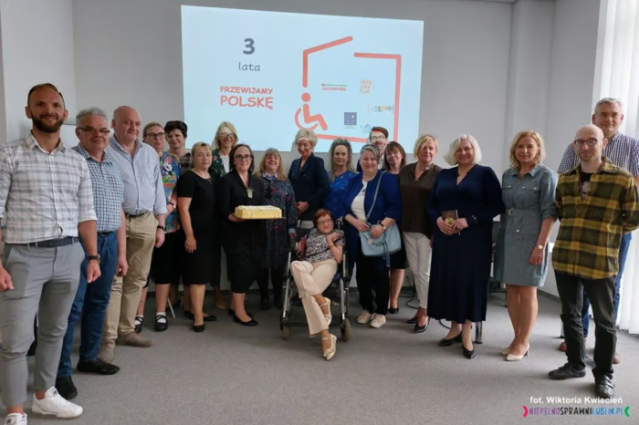 Pokaż zdjęcie: Do pamiątkowego zdjęcia ustawiła się grupa 19 osób. Przed grupą osób na wózku inwalidzkim siedzi kobieta. Na ścianie slajd z logo akcji.