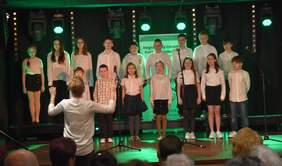 Pokaż zdjęcie: Grupa dzieci i młodzieży w białych koszulach i czarnych spódnicach lub spodniach śpiewa na scenie podczas obchodów Światowego Dnia Inwalidy w Białymstoku. Dyrygentka w białej bluzce prowadzi chór, stojąc tyłem do publiczności. W tle widoczne są reflektory sceniczne oraz baner Regionalnego Ośrodka Polityki Społecznej w Białymstoku.