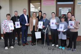 Pokaż zdjęcie: Grupa dziesięciu osób w strojach codziennych i półformalnych stoi przed wejściem do budynku. Kilka osób prezentuje otrzymane dyplomy z okrągłą grafiką.