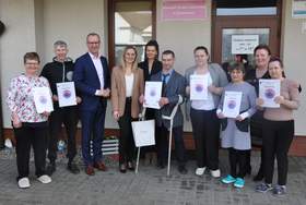 Grupa dziesięciu osób w strojach codziennych i półformalnych stoi przed wejściem do budynku. Kilka osób prezentuje otrzymane dyplomy z okrągłą grafiką.