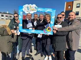 Pokaż zdjęcie: Na ulicy stoi grupa spacerowiczów. Trzymają w dłoniach niebieską ramkę, na której napisana jest nazwa organizacji "Siła Małych Kroków" i symbol nieskończoności w różnobarwnych kolorach. Widać błękit nieba. 