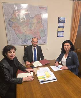 Pokaż zdjęcie: Dwie kobiety oraz mężczyzna siedzą przy owalnym stole i rozmawiają.