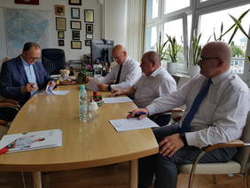 widok z boku, z lewej strony jeden mężczyzna w garniturze, z prawej trzej w białych koszulach siedzą przy stole, podpisują dokumenty