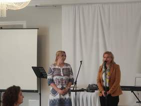 Pokaż zdjęcie: Na scenie przy mikrofonach stoją dwie kobiety, jedna z nich przemawia, w tle widać ekran do prezentacji