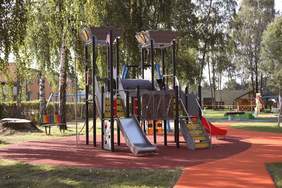 Pokaż zdjęcie: Na środku placu zabaw znajduje się wieża ze zjeżdżalniami, drabinkami, mini ścianką wspinaczkową. W tle widać drzewa. 