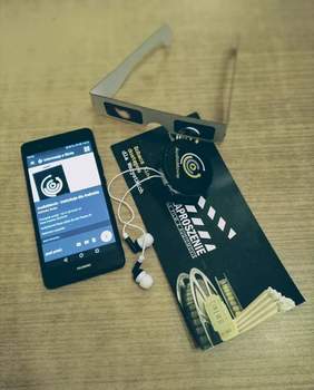 Pokaż zdjęcie: Ułożone obok siebie przedmioty: telefon z uruchomioną aplikacją AudioMovie, zaproszenie na seans, słuchawki wystające z okrągłego pudełeczka oraz okulary symulujące niepełnosprawność wzroku