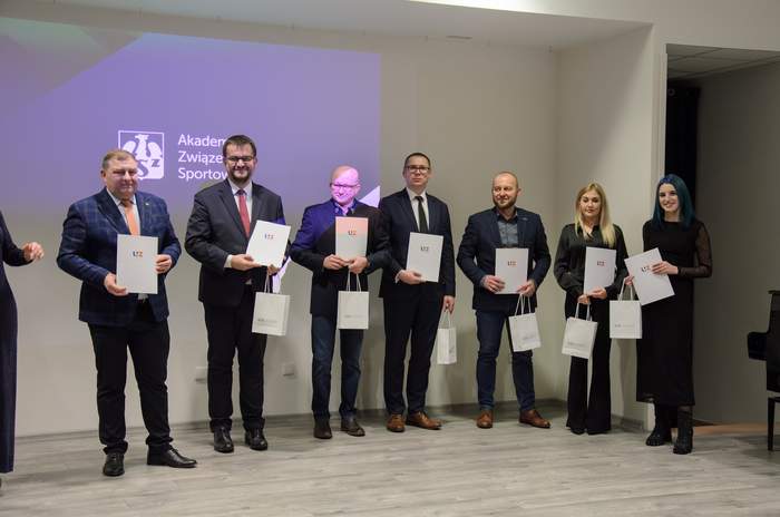 Pokaż zdjęcie: Goście i laureaci spotkania, ubrani na galowo, stoją obok siebie na scenie i trzmają w rękach dyplomy i torebki upominkowe