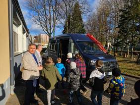 Pokaż zdjęcie: Grupa dzieci i dwóch mężczyzn stoi przy nowoczesnym, ciemnym busie ozdobionym czerwoną kokardą, zaparkowanym obok budynku w otoczeniu drzew. Dzieci z ciekawością podchodzą do pojazdu.