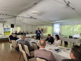 Pokaż zdjęcie: Mężczyzna w granatowej marynarce i białej koszuli trzyma mikrofon w obu rękach i mówi do osób siedzących za długim stołem. Wszyscy ubrani są na galowo. Wszystko znajduje się w białym elegancji namiocie. Do dokoła zieleń.