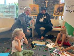 Pokaż zdjęcie: dwóch mężczyzn rozmawia, na pierwszym planie dwójka dzieci malujących nogami. Z tyłu wyeksponowane trzy obrazy przedstawiajace wierzby o zachodzie słońca