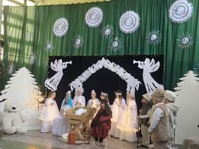 Pokaż zdjęcie: Przy żłóbku z dzieciątkiem Jezus zebrali się Maryja z Józefem, pastuszkowie oraz aniołki. W tle świąteczna dekoracja.