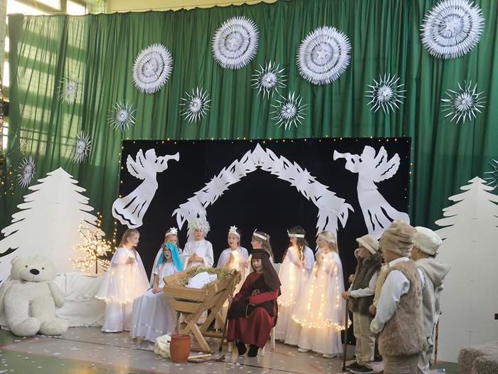 Pokaż zdjęcie: Przy żłóbku z dzieciątkiem Jezus zebrali się Maryja z Józefem, pastuszkowie oraz aniołki. W tle świąteczna dekoracja.