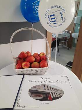 Pokaż zdjęcie: Na stoliku leży pamiątkowa księga. Napisany jest na niej tekst: Otwarcie Powiatowego Centrum Zdrowia. Za nią stoi kosz z jabłkami oraz białe i niebieskie balony. 