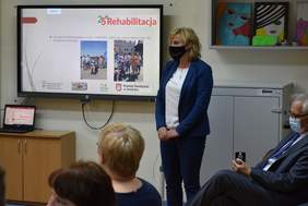 Pokaż zdjęcie: Kobieta stoi przed ekranem na którym jest podsumowanie programu Rehabilitacja 25 plus         Rehabilitacja 2