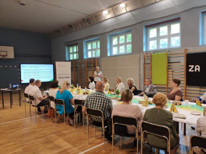 Pokaż zdjęcie: W sali gimnastycznej ustawiony jest długi stół przy którym siedzą ludzie na ekranie wyświetlona jest prezentacja obok stoi kobieta z mikrofonem i baner z logo PFRON