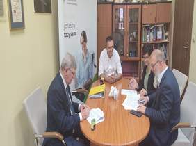 Pokaż zdjęcie: pomieszczenie w Oddziale łodzkim po środku stoi duży owalny stół przy nim siedzą cztery osoby i podpisują dokumenty w tle baner z logo PFRON