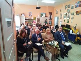 Pokaż zdjęcie: W sali domu opieki na krzesłach siedzą zaproszeni goście, w pierwszym rzędzie mężczyźni w garniturach oraz kobieta w bezowej sukience trzymająca kwiaty