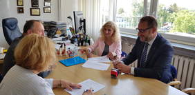 4 osoby podpisujące umowę siedzące przy stole, z lewej kobieta (widok z tyłu) i fragment twarzy mężczyzny, z prawej strony uśmiechnięta kobieta w marynarce i uśmiechnięty mężczyzna w garniturze