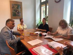 Pokaż zdjęcie: Przy stole siedzą trzy kobiety i mężczyzna. Podpisują dokumenty. Mężczyzna Trzyma w ręku pieczątkę. W tle okno, parapet i stojące nas nim kwiaty.