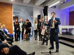 Pokaż zdjęcie: Na scenie stoi osiem osób, mężczyzna w garniturze stoi przy mikrofonie składa życzenia, osoby stojące za nim trzymają w rękach prezenty.