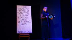 Pokaż zdjęcie: Na scenie stoi plakat XIX Biłgorajski Przegląd Twórczości Muzyczno -Teatralnej Osób Niepełnosprawnych, obok plakatu kobieta ubrana w ciemną sukienkę