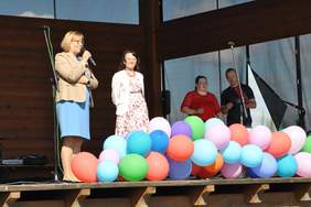 Pokaż zdjęcie: Drewniana scena ozdobiona kolorowymi balonami. Na scenie stoją cztery osoby. Kobieta przemawia do mikrofonu.