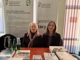 Pokaż zdjęcie: Dwie kobiety siedzą przy stole promocyjnym z materiałami PFRON i CIDON; na blacie ułożone broszury, notesy, długopisy i butelki wody, a za nimi stoją roll-upy z informacjami o wsparciu dla osób z niepełnosprawnościami.