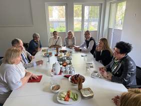 Pokaż zdjęcie: Grupa osób siedzi przy białym stole, rozmawia przy kawie i przekąskach. Na stole owoce, warzywa, ciastka, dzbanek z kawą i filiżanki. Spotkanie odbywa się w jasnym pomieszczeniu z dużymi oknami.