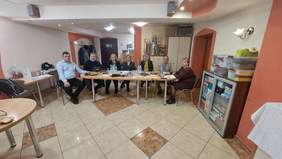 Pokaż zdjęcie: Siedem osób siedzi przy stole, wszyscy mają twarz zwróconą do aparatu. Znajdują się w pomieszczeniu, za nimi szafa oraz komoda i drzwi wyjściowe.